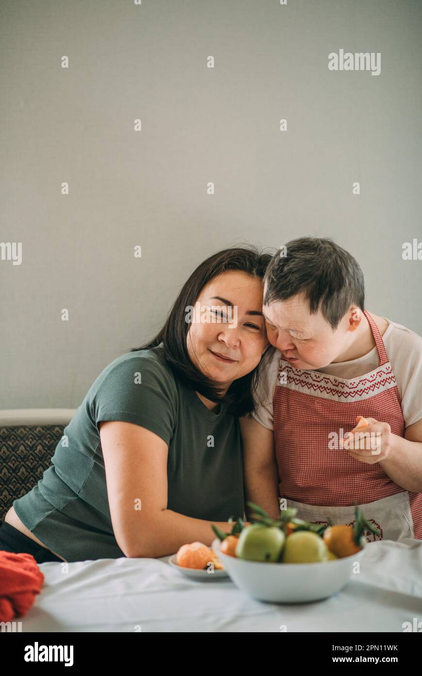 An elderly woman with down syndrome gently leans against an Asian woman