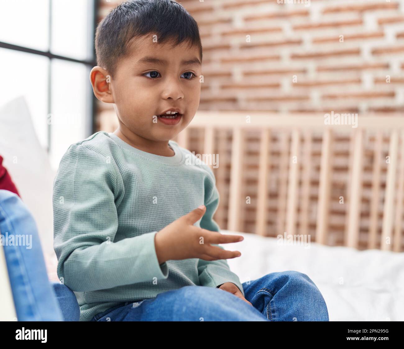 Adorable hispanic boy sitting on bed with relaxed expression at bedroom