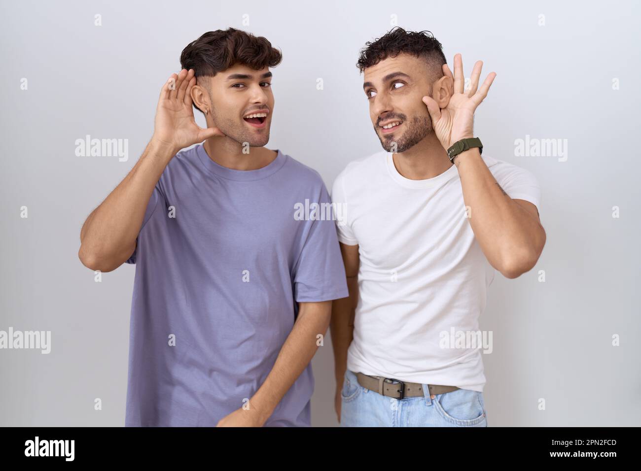 Homosexual gay couple standing over white background smiling with hand over ear listening an