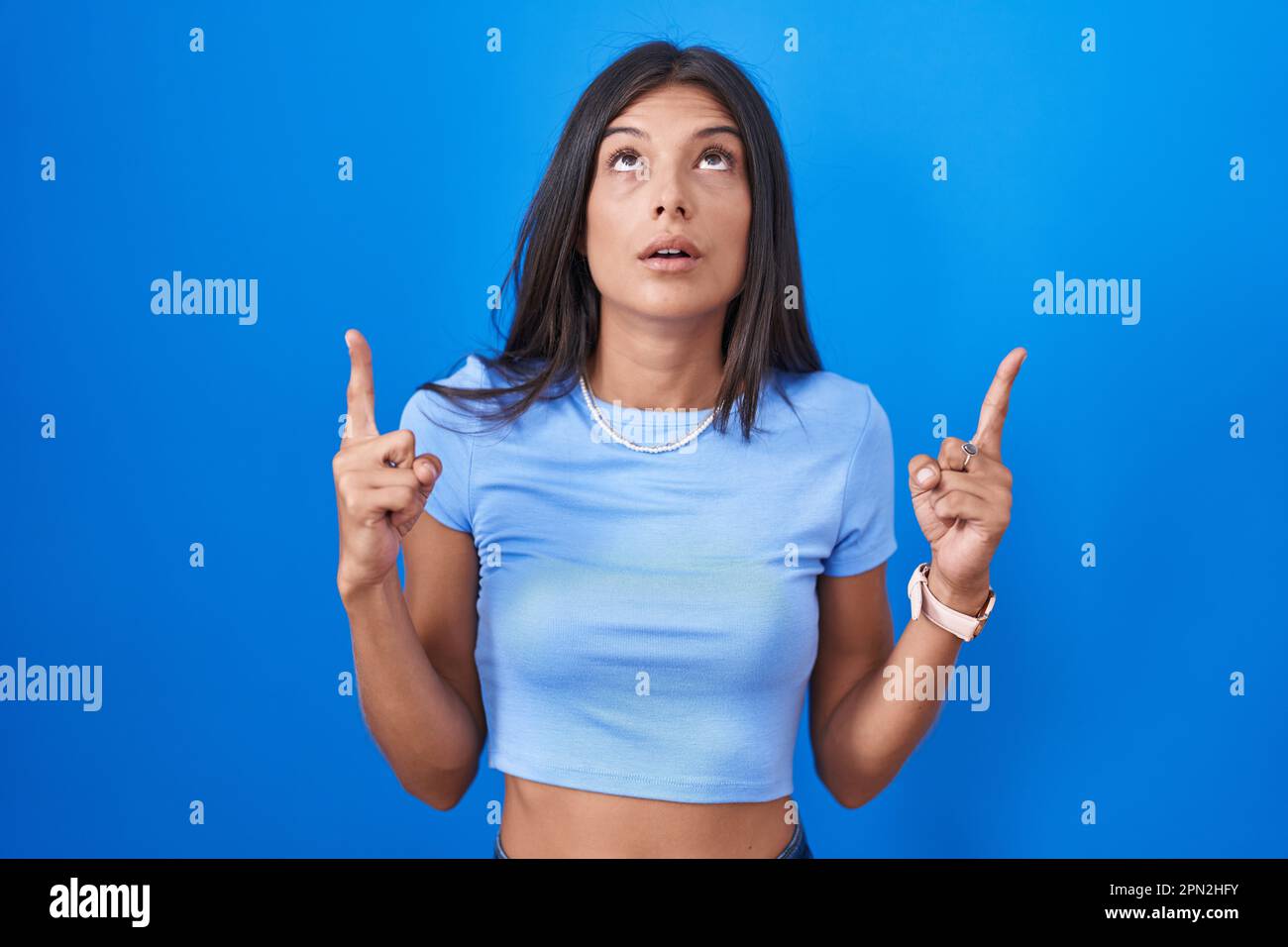 Brunette young woman standing over blue background amazed and surprised ...