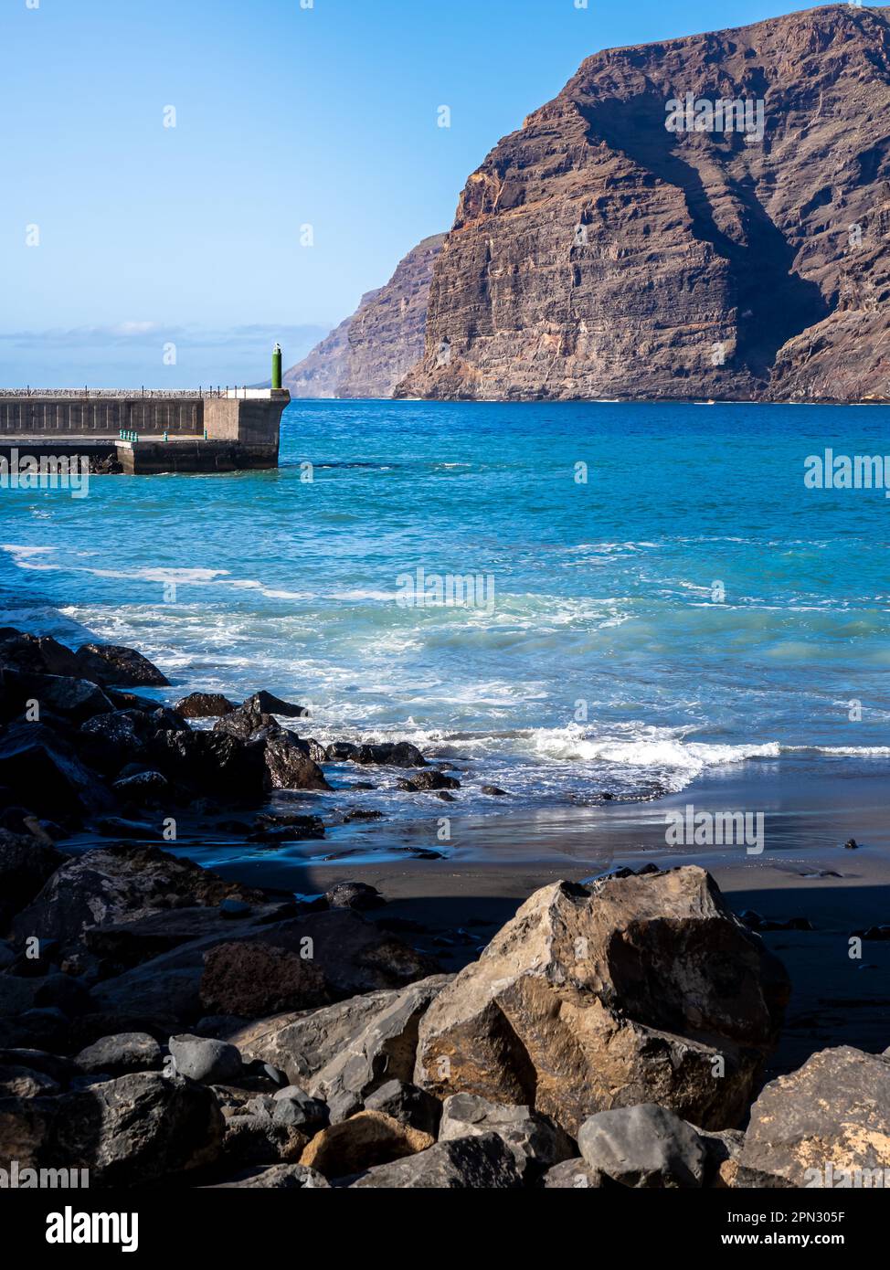 Sunlight on the majestic Los Gigantes cliffs and the black sand of Playa Los Guíos beach, with Los Gigantes harbor entrance and rugged rocks. Stock Photo