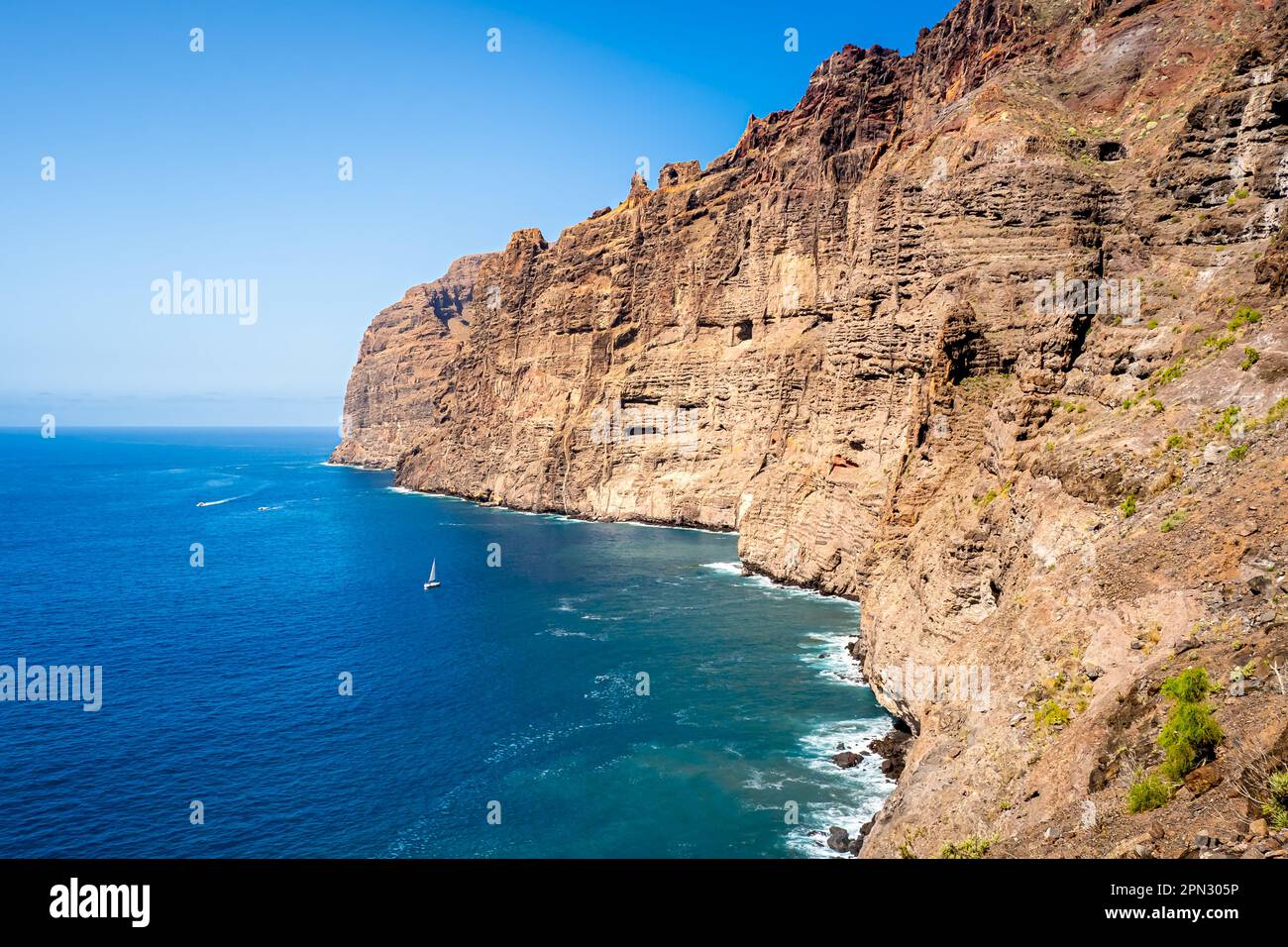 Los Gigantes cliffs tower above the rocky coastline of Tenerife's western shore while a small sailboat sails through the calm waters. Stock Photo