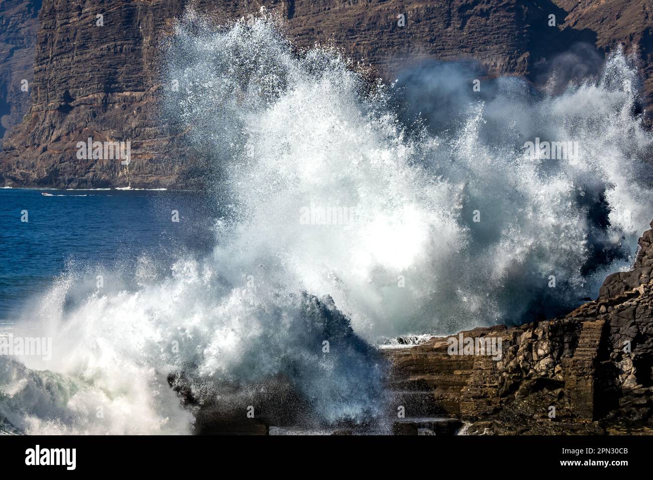 The mighty ocean sends a massive wave crashing against the rocky shore, creating a spectacular display of natural power at Acantilados de los Gigantes Stock Photo