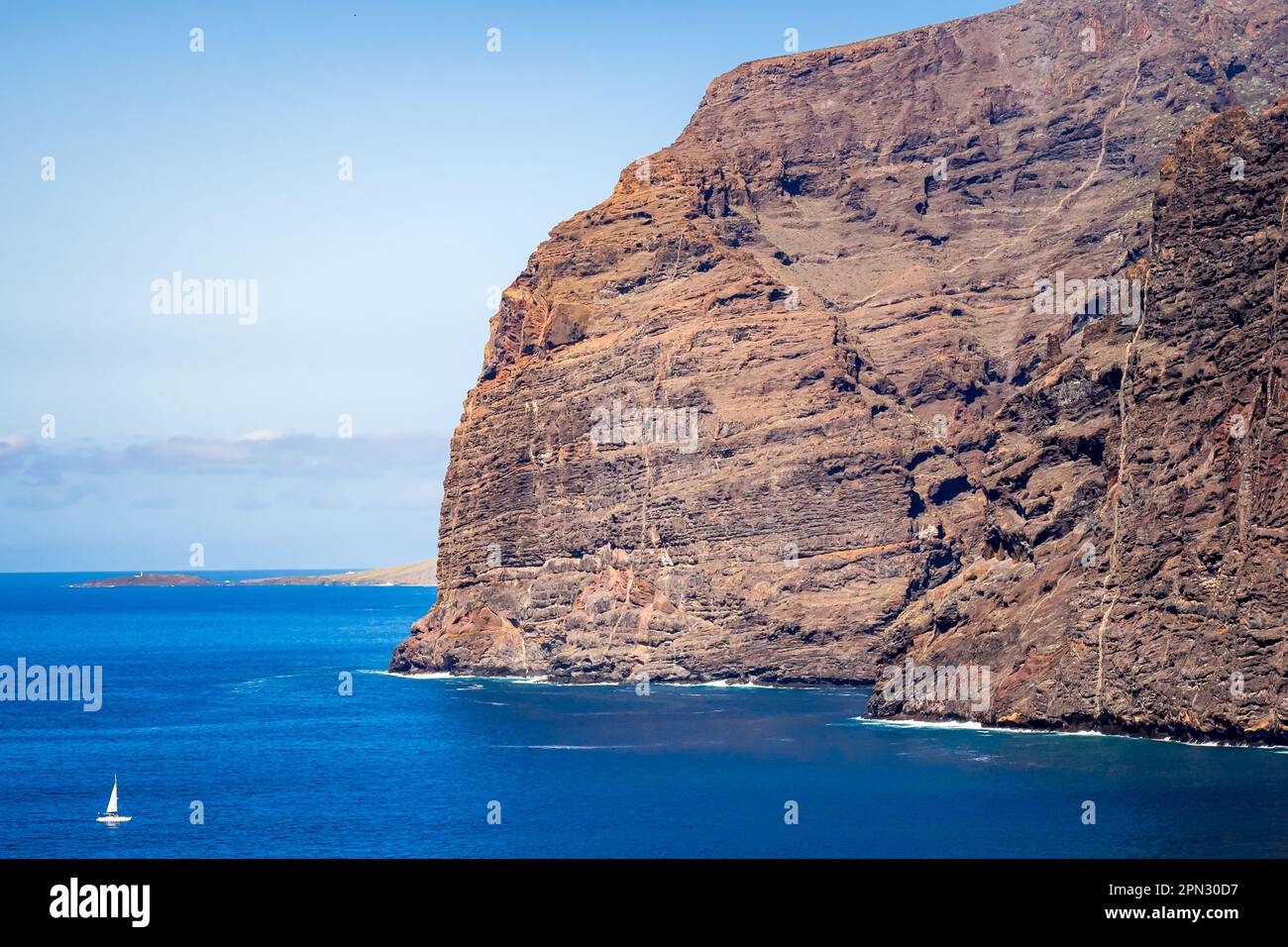 Majestic Acantilados de Los Gigantes cliffs standing tall, while a small sailing boat sails by, with Punta de Teno headland and Teno lighthouse. Stock Photo