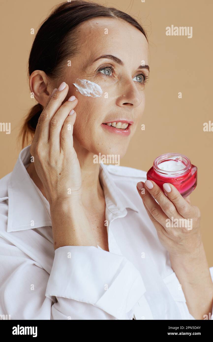 Smiling Modern Woman With Facial Cream Jar In White Shirt Against Beige