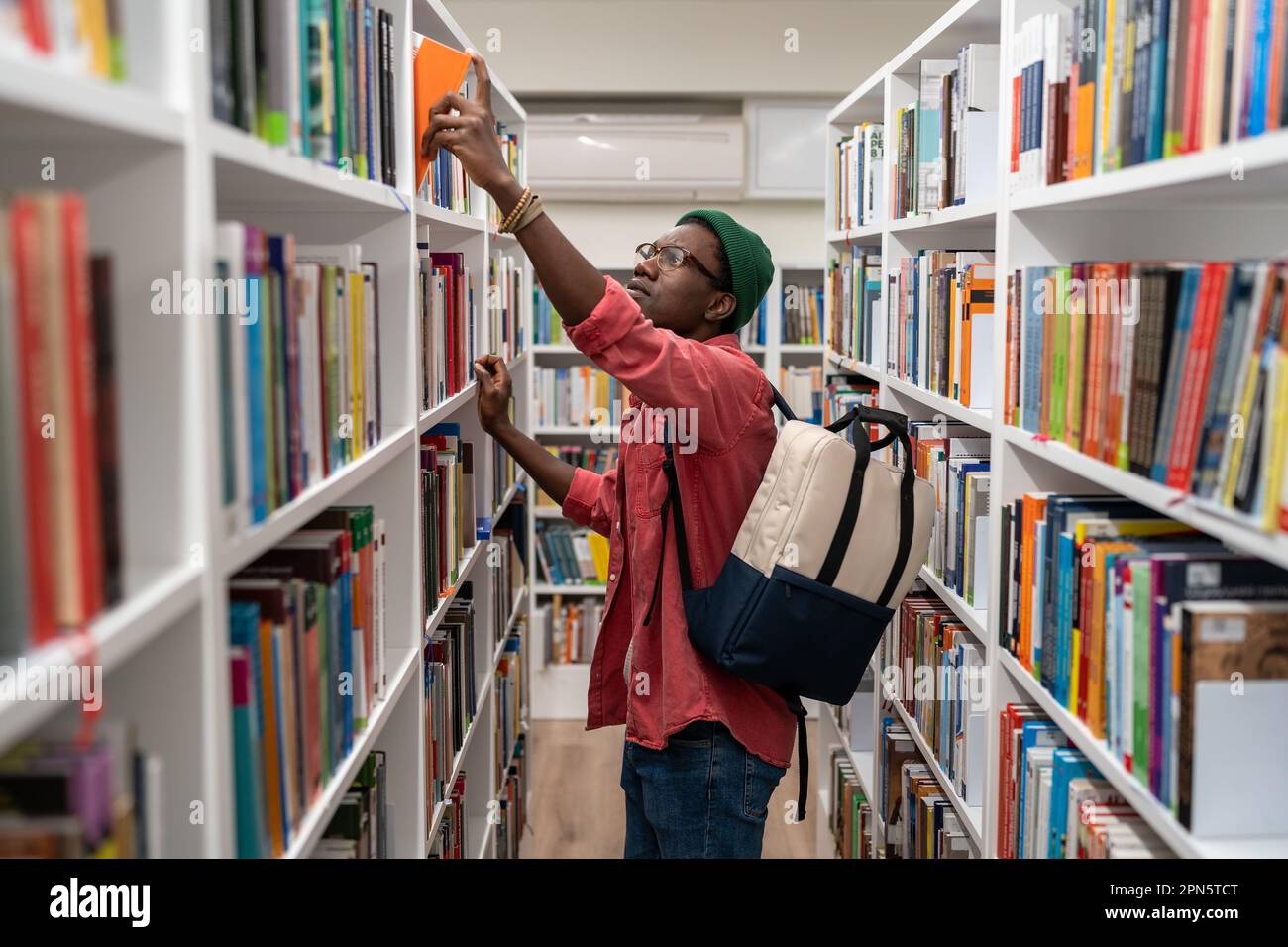 Nerd student african american man choosing book in university library taking it from shelf. Stock Photo
