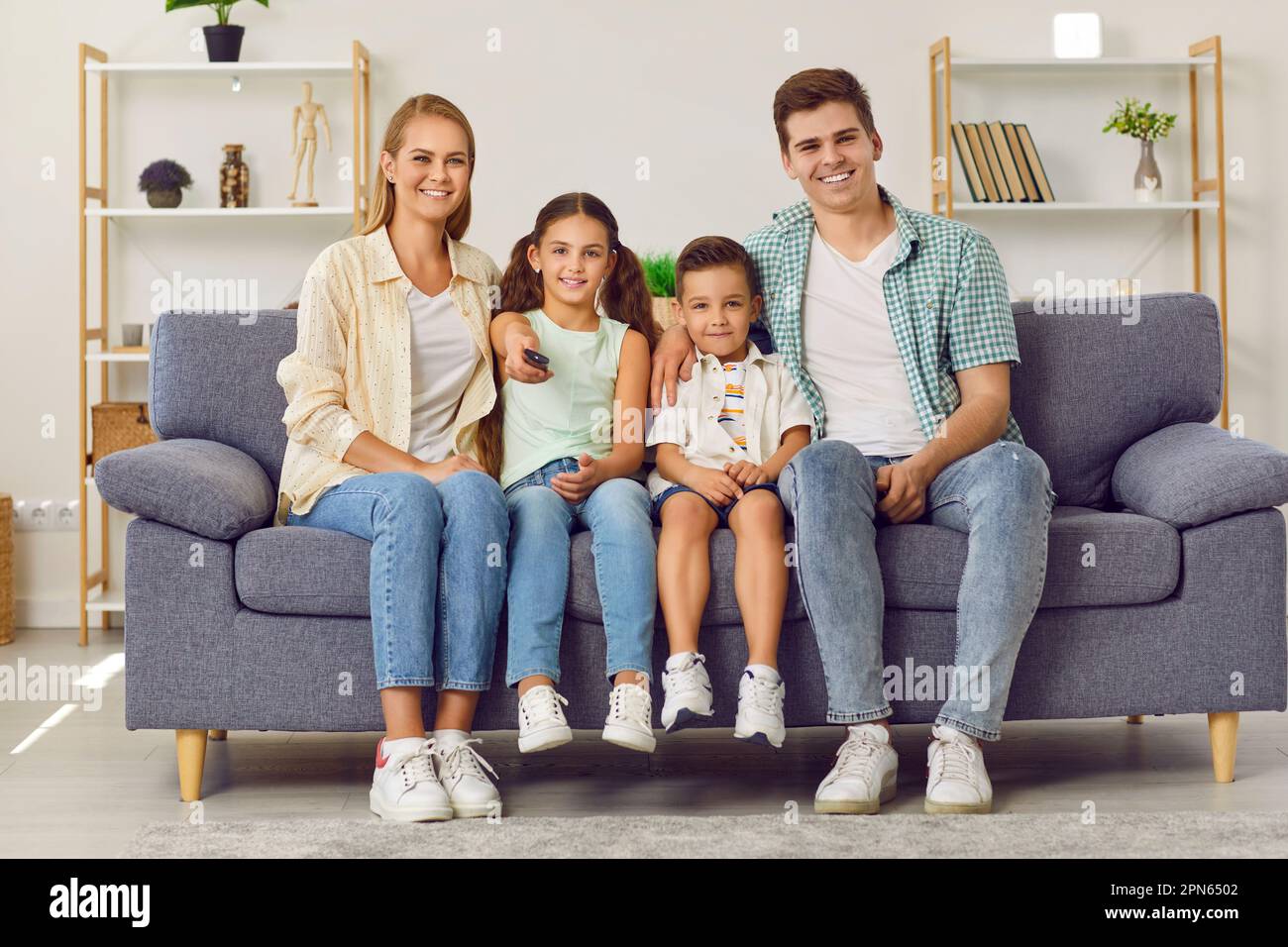 Young happy Caucasian family with two children watching TV together sitting on sofa in living room. Stock Photo