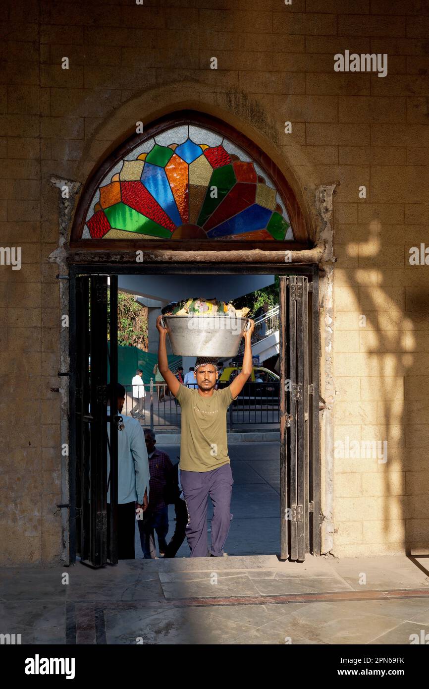 A porter carrying goods on his head enters Chhatrapati Shivaji Maharaj Terminus in Mumbai, India, through a side entrance headed by a colorful mosaic Stock Photo