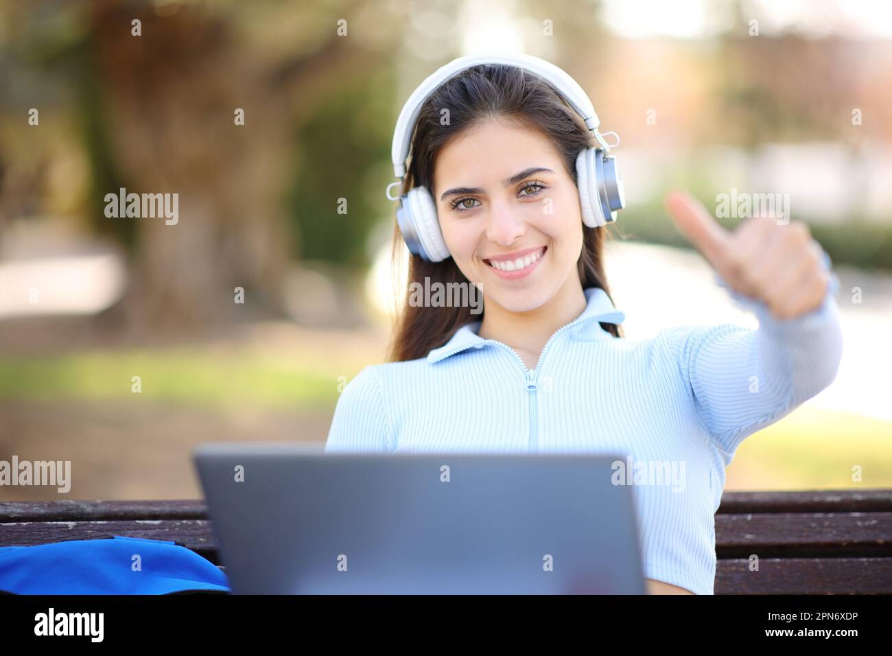 Happy student with laptop and headphone with thumbs up on a bench in a park Stock Photo