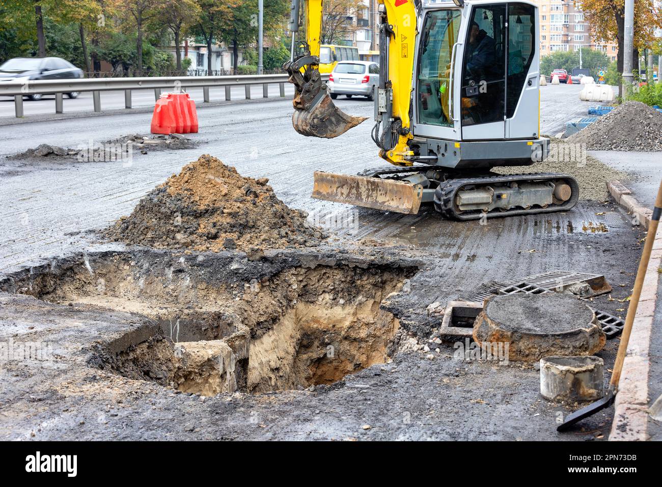 A construction worker on a mobile road excavator repairs sewer storm hatches on a fenced-off roadway of a city street on a summer day. Stock Photo