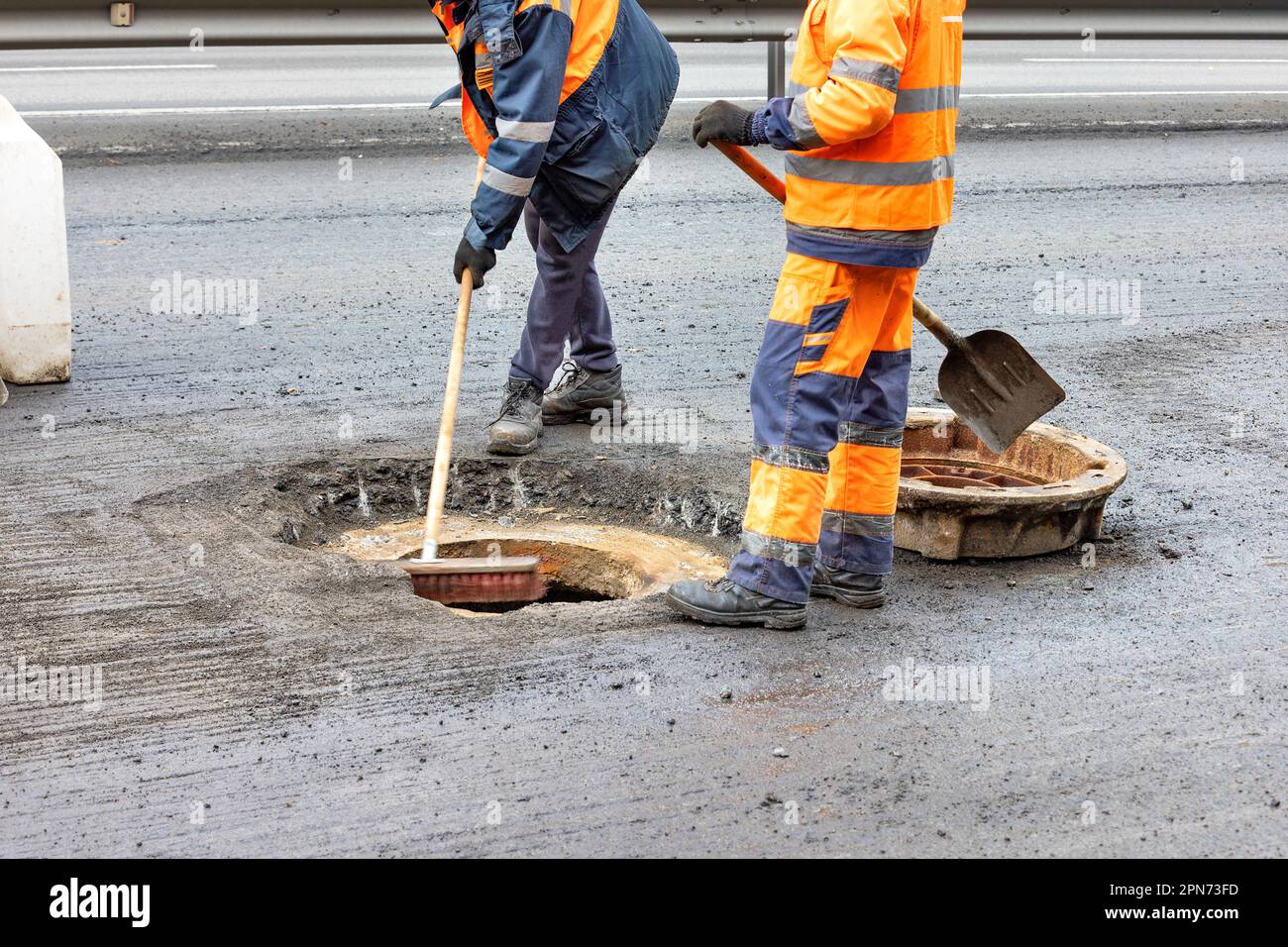 A team of road workers in orange-and-blue overalls with a shovel and brush clear a site for a new sewer manhole on the roadway. Copy space. Stock Photo