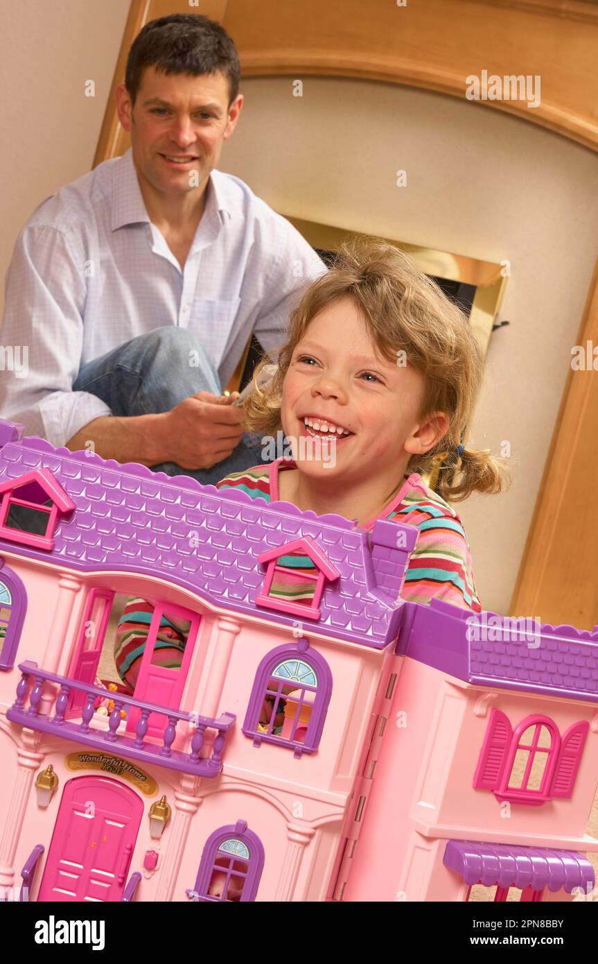 Young girl playing with a pink dolls house with her father in the background Stock Photo