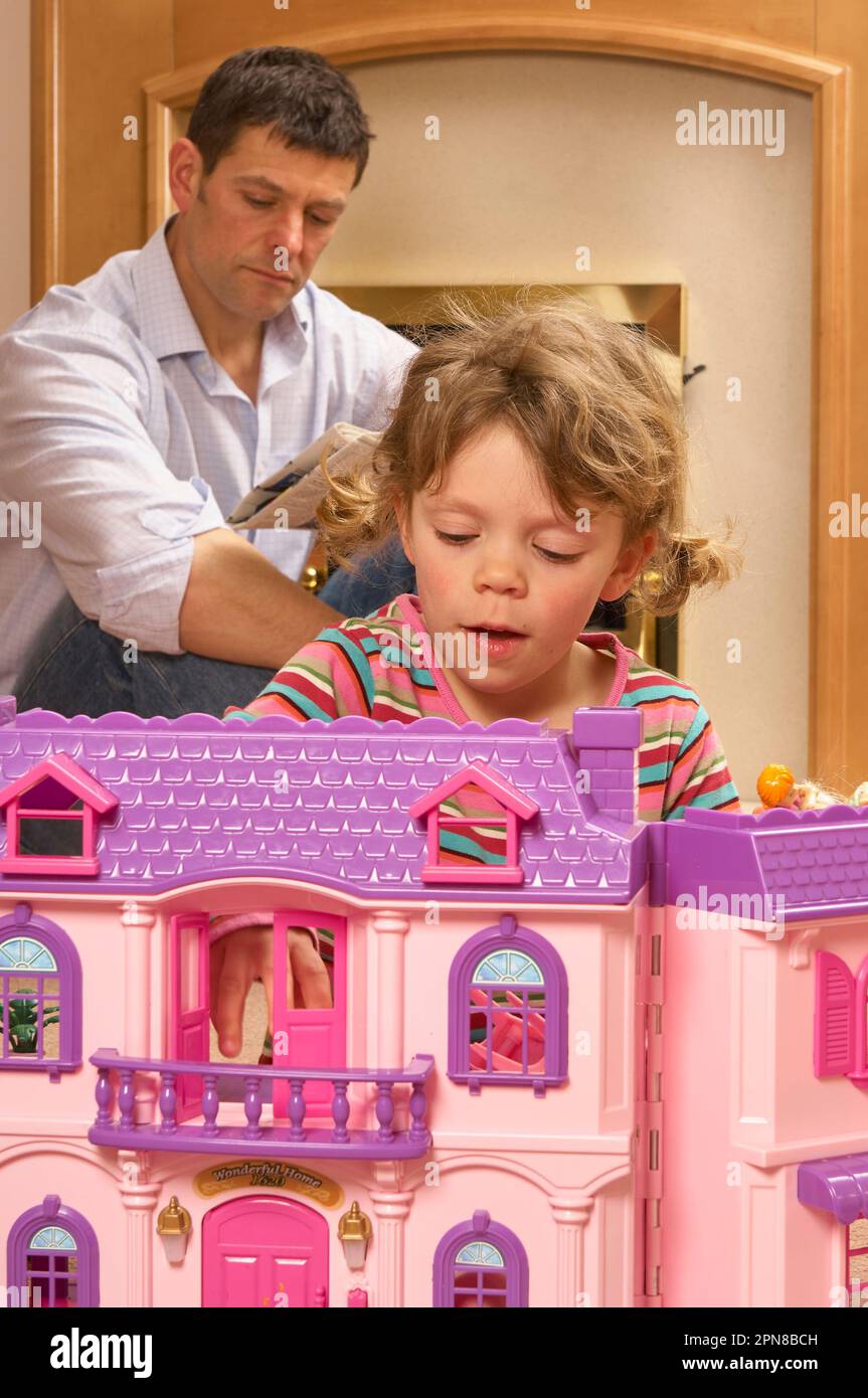 Young girl playing with a pink dolls house with her father in the background Stock Photo