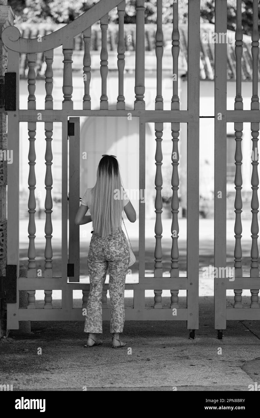 A grayscale of a young female stands among a white picket fence, opening the gate Stock Photo