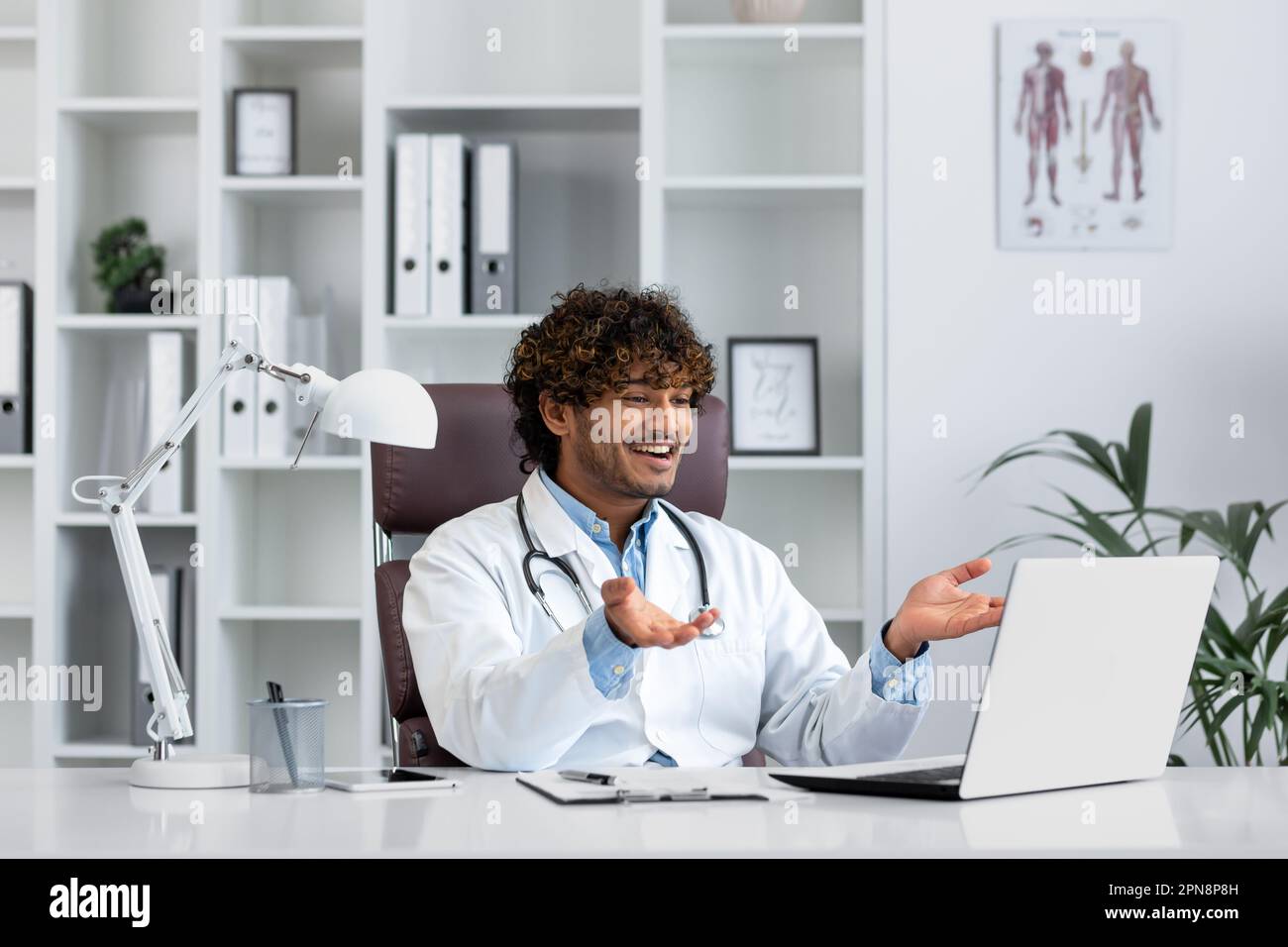 Hindu doctor consulting patients remotely, man working inside modern clinic office, using laptop for online video call, talking and smiling friendly to patient. Stock Photo