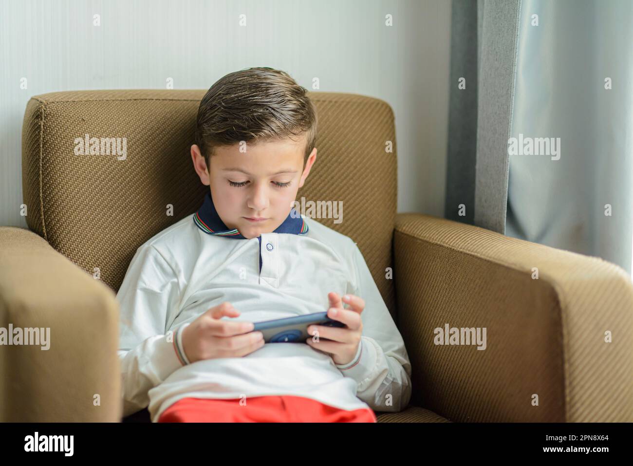 Child using smartphone while sitting on a chair. Technology use by children and its consequences, including poor posture. Stock Photo