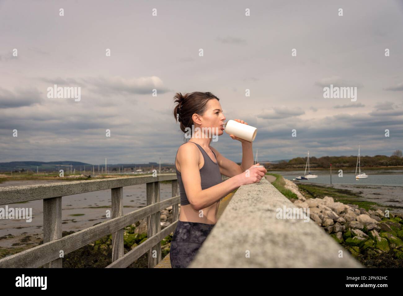sportswoman drinking water from bottle after workout, resting on a bridge by the coast Stock Photo