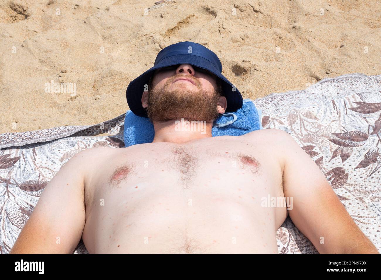 a large man in a Panama hat with white skin and a red beard is sunbathing on the beach on a sunny day. Rest and relaxation. Stock Photo