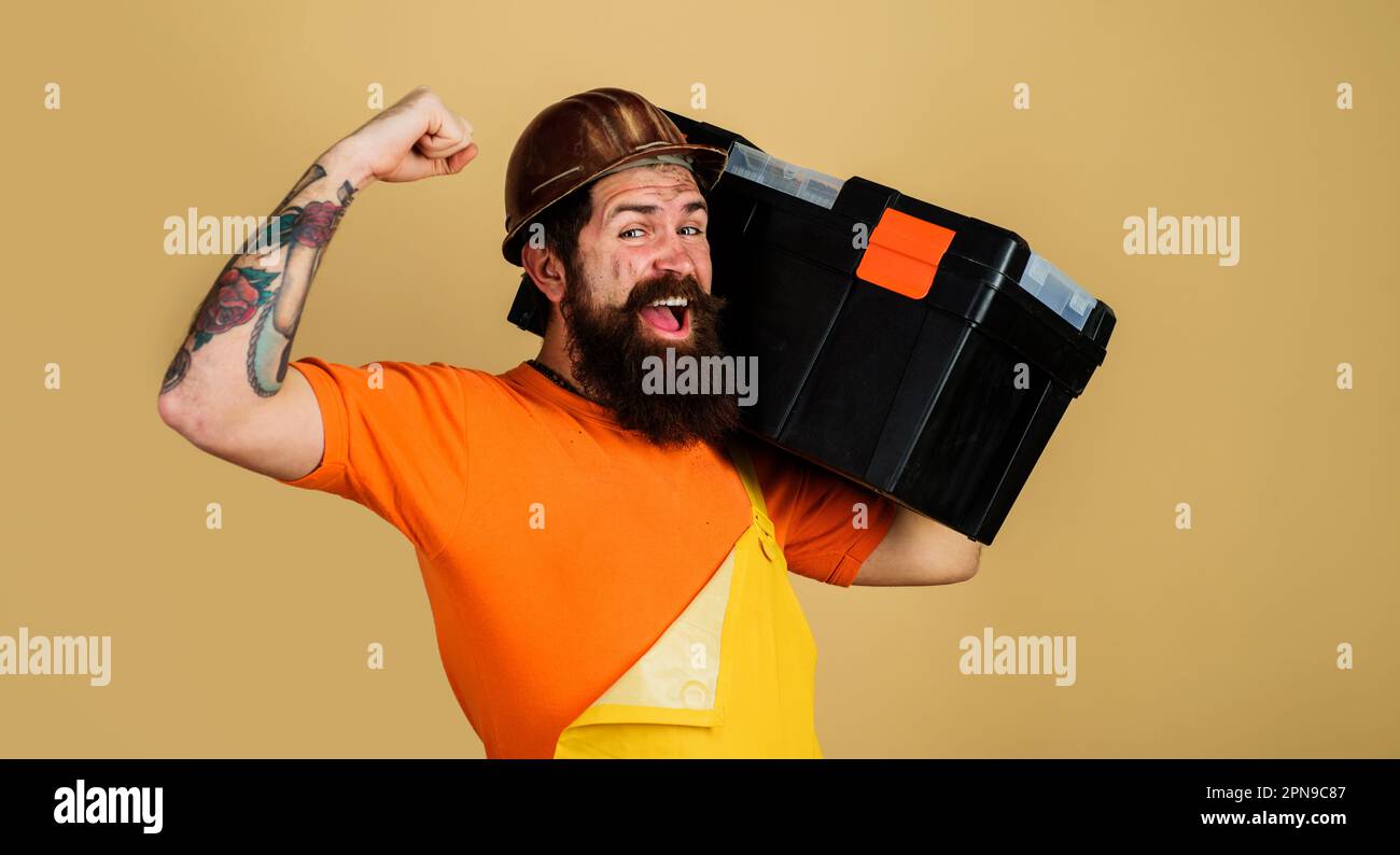 Happy bearded man in builder uniform with toolbox. Handyman with repair kit. Repairman in overalls carry toolbox. Male builder in hardhat with toolbox Stock Photo