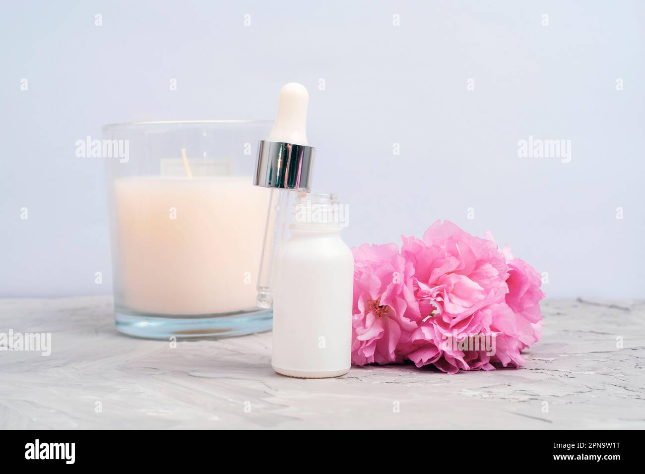 White serum dropper bottle, pink cherry blossom and aroma candle on concrete background. Closeup, front view. Stock Photo
