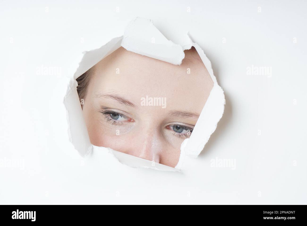 young woman peeking through hole torn in paper wall Stock Photo