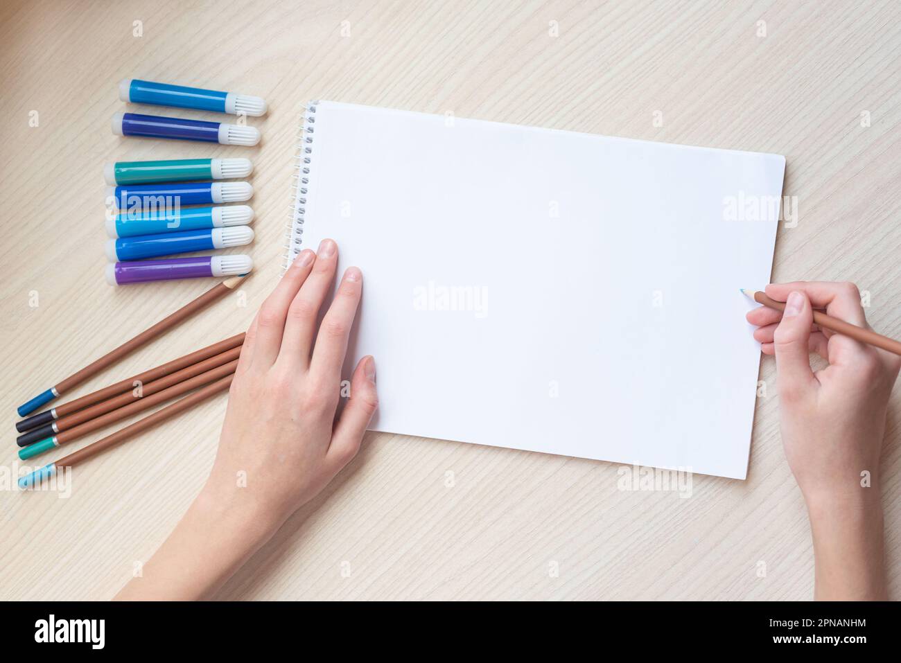 A clean white sheet of paper on the table, the girl's hands holding a pencil. Mockup, top view. Stock Photo