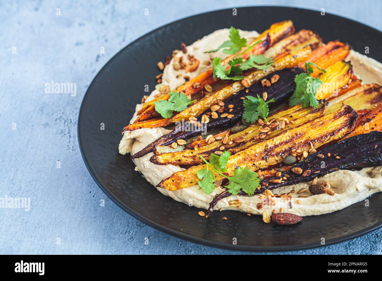 Multicolored baked carrots with hummus on a black plate. Vegan recipe. Stock Photo