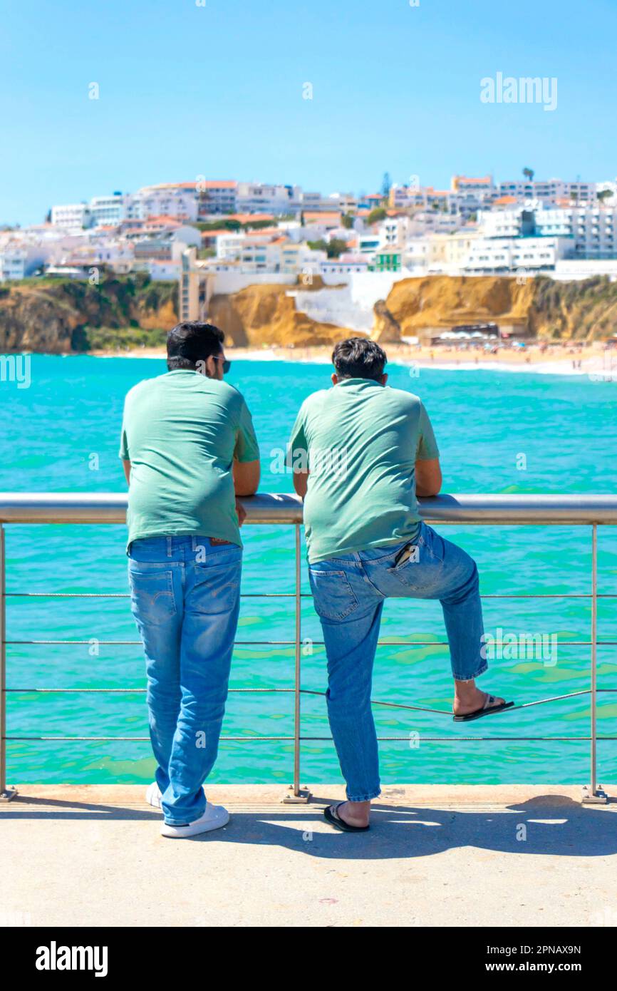 two asian men in matching clothes look out to sea from the pier in albufeira algarve portugal Stock Photo