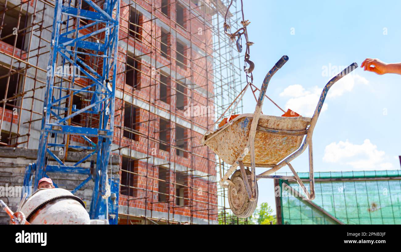 Crane is carrying wheelbarrow full of red bricks, pulled up above building site, keeping balance and direction, in the background is edifice under con Stock Photo