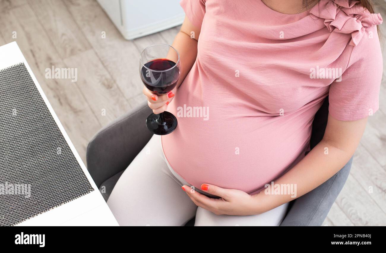 A pregnant girl sits in the kitchen at the table and drinks alcohol. Bad habit during pregnancy. Stock Photo