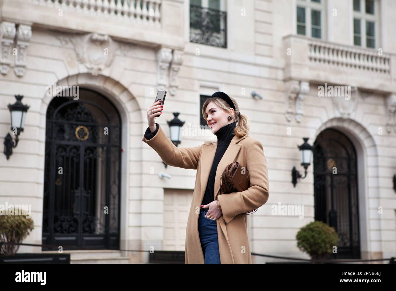 Beautiful young happy woman tourist takes selfie against beautiful architecture building while traveling on historic city street. Fashionable female m Stock Photo