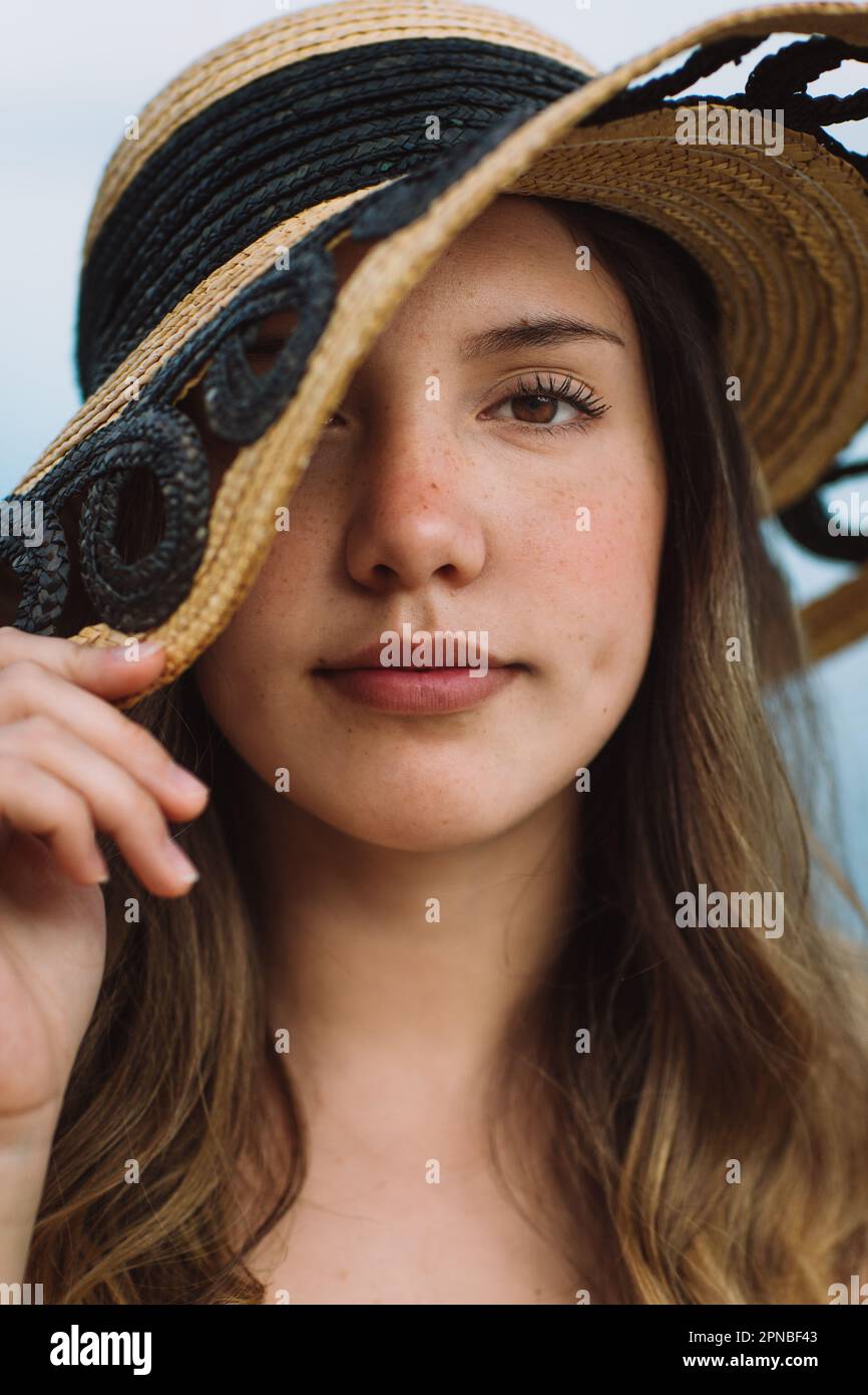 Portrait of female model covering face with straw hat and looking at camera while resting on summer vacation against blue background Stock Photo