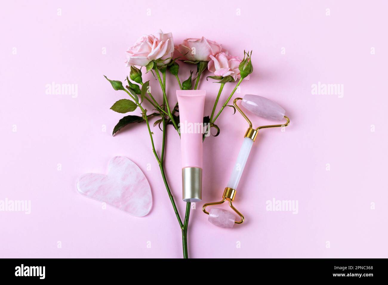 Pink jade roller and gua sha for face massage with cream or gel for the face and flower roses. Facial massager tools. Anti age, lifting and toning car Stock Photo