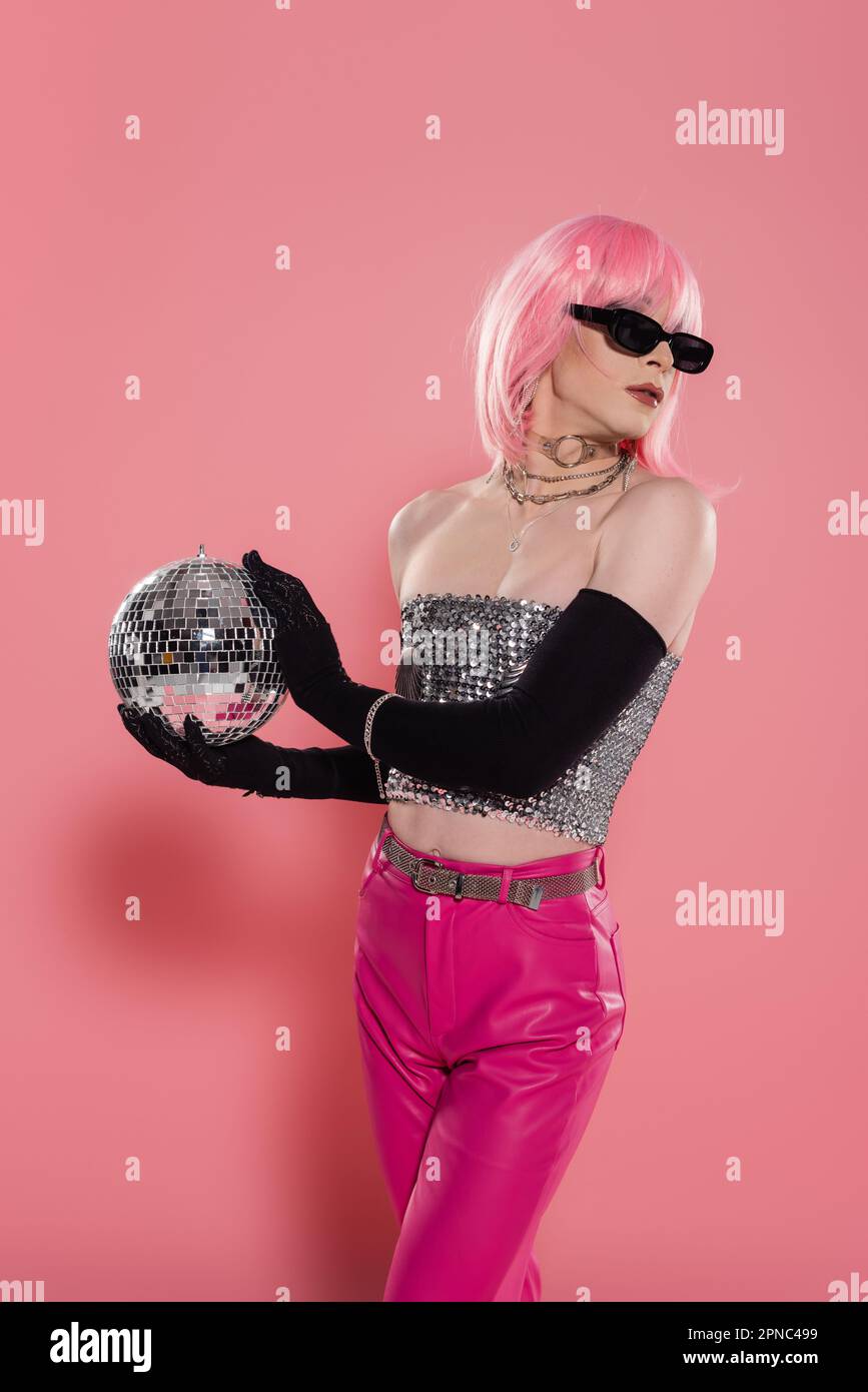 Fashionable drag queen in sunglasses and silver top holding disco ball on pink background,stock image Stock Photo