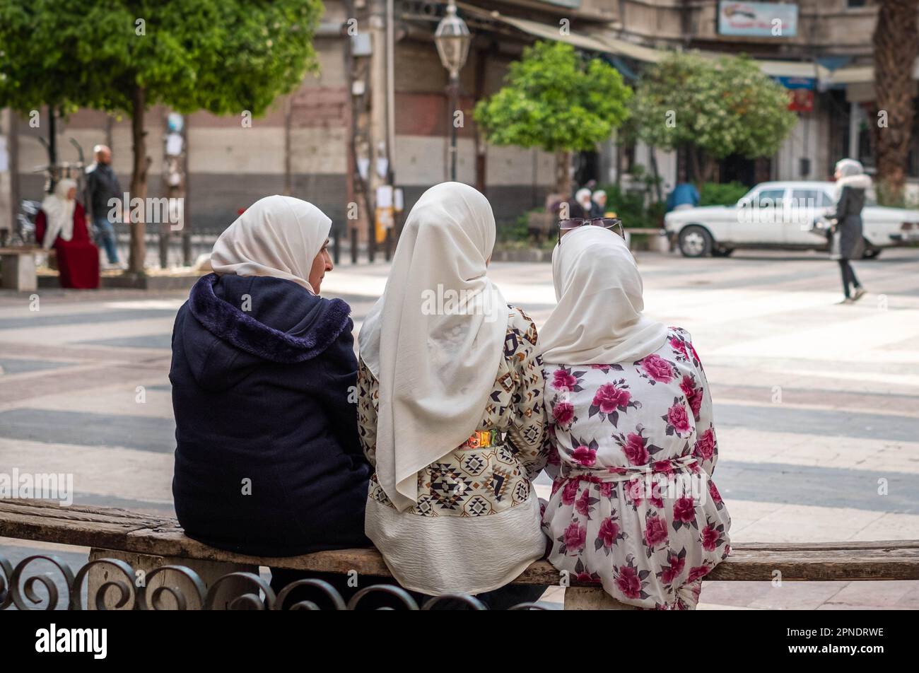 Three muslim women or girls wearing headscarf / hijab sitting on bench in the street Stock Photo