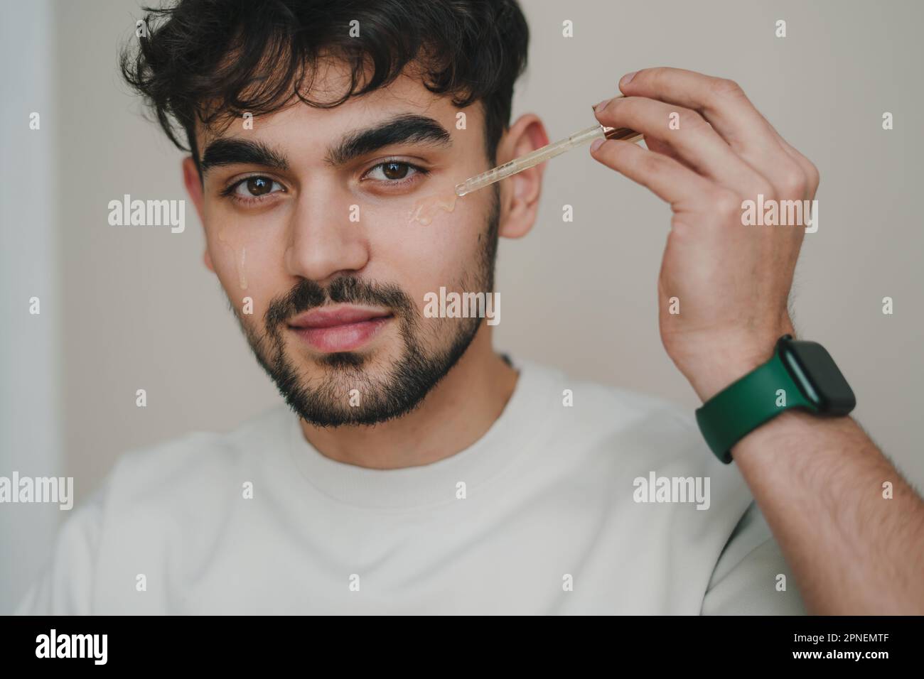 Portrait of a man applying oil serum on his face at bathroom looking at camera. Self care