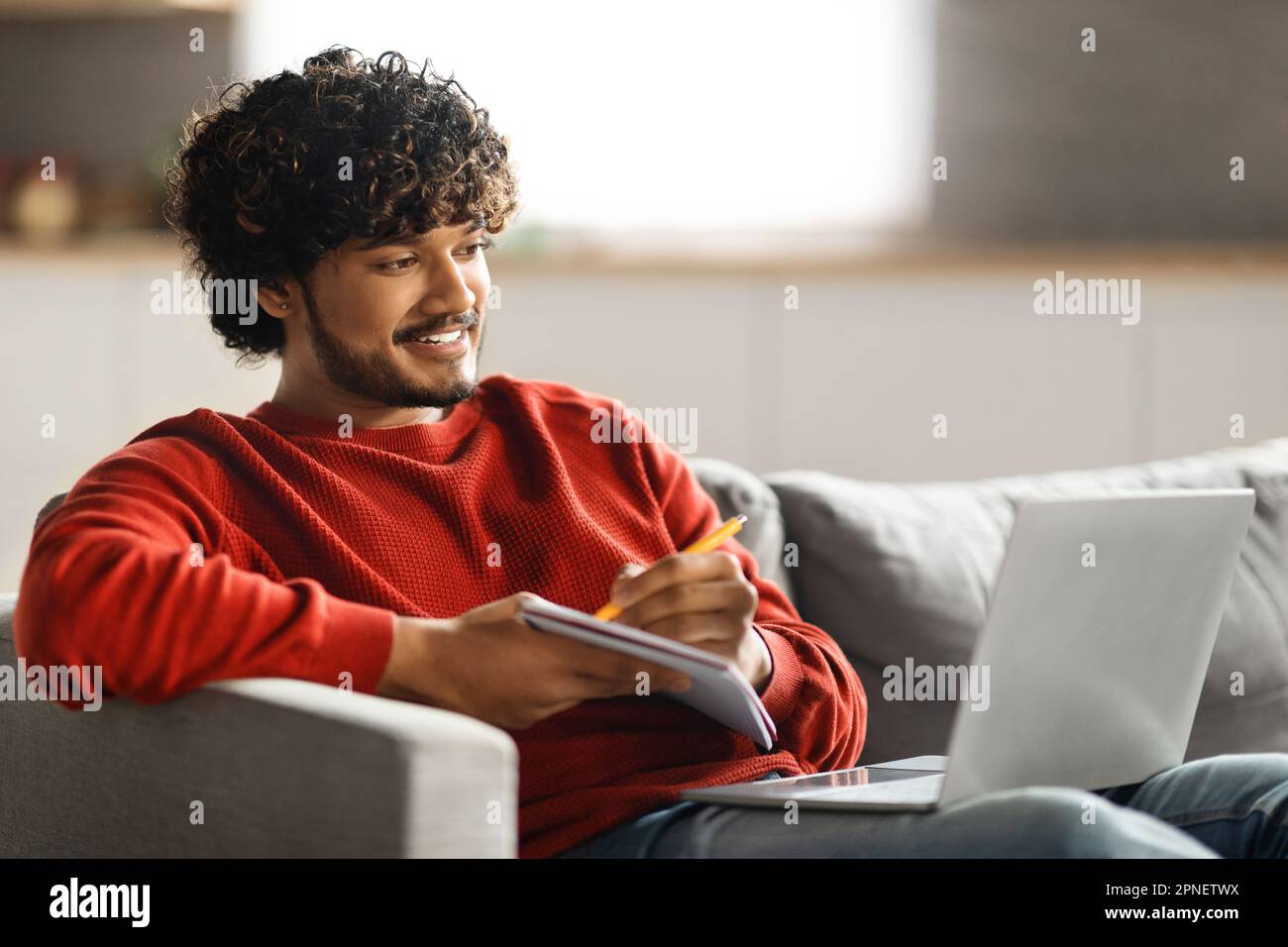 Online Education. Handsome young indian man using laptop and taking notes Stock Photo