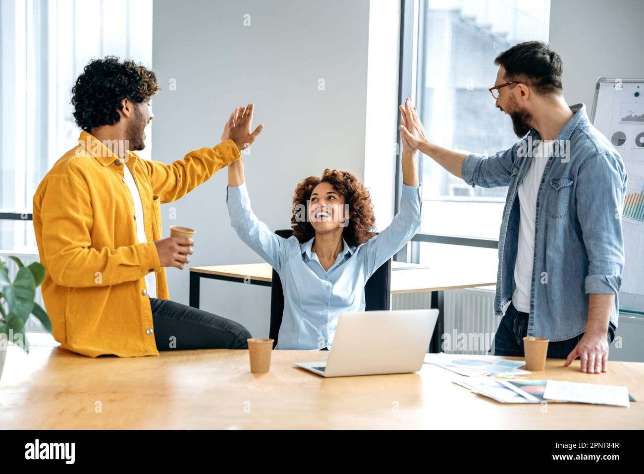 Cheerful colleagues, multiracial team, sitting in a creative office, working on a new project, a startup, developing a strategy and deadlines, happy with the result, celebrate success, achievement Stock Photo