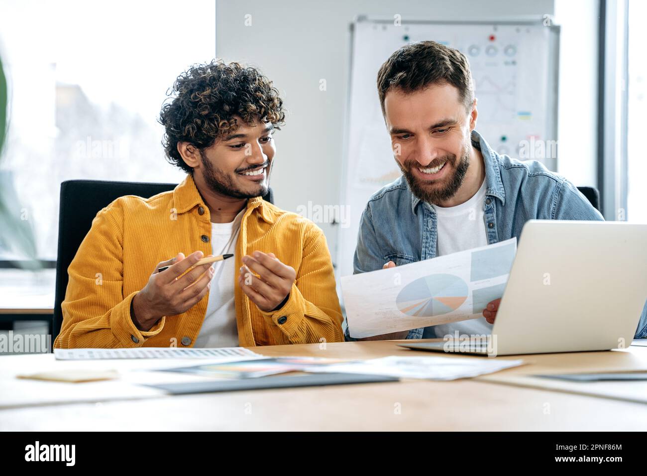 Two joyful male multiracial employees sit in the office at the table ...