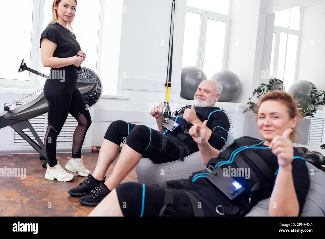 https://c8.alamy.com/comp/2PNH4XA/happy-aged-couple-in-ems-suits-sits-on-massage-chairs-after-electrical-muscle-stimulation-workout-with-personal-coach-in-gym-or-fitness-club-smiling-2PNH4XA.jpg