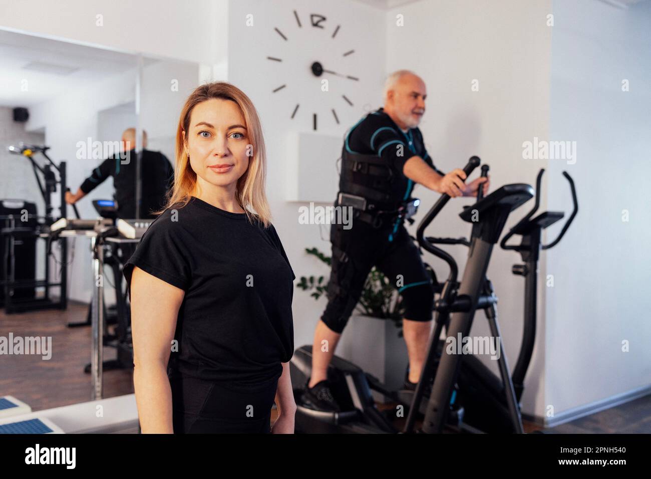Personal female trainer with her client in ems suit in gym. Sports senior grayhaired man is exercising on elliptical trainer on background. Electrical Stock Photo