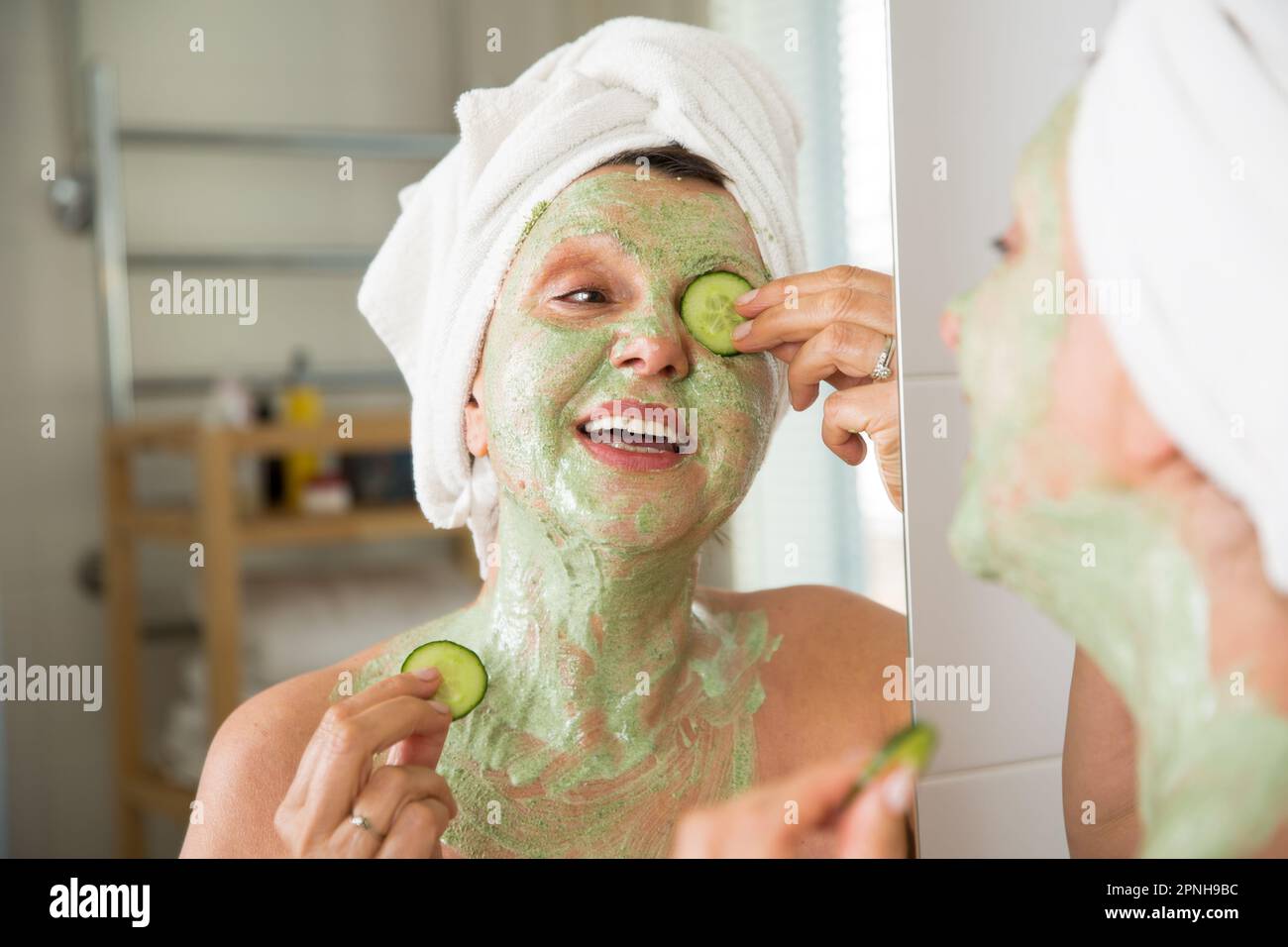 Mature beautiful woman applying green homemade DIY facial mask on skin. Looking in the mirror in bathroom, having fun. Wrapped in a towel. Skincare Stock Photo