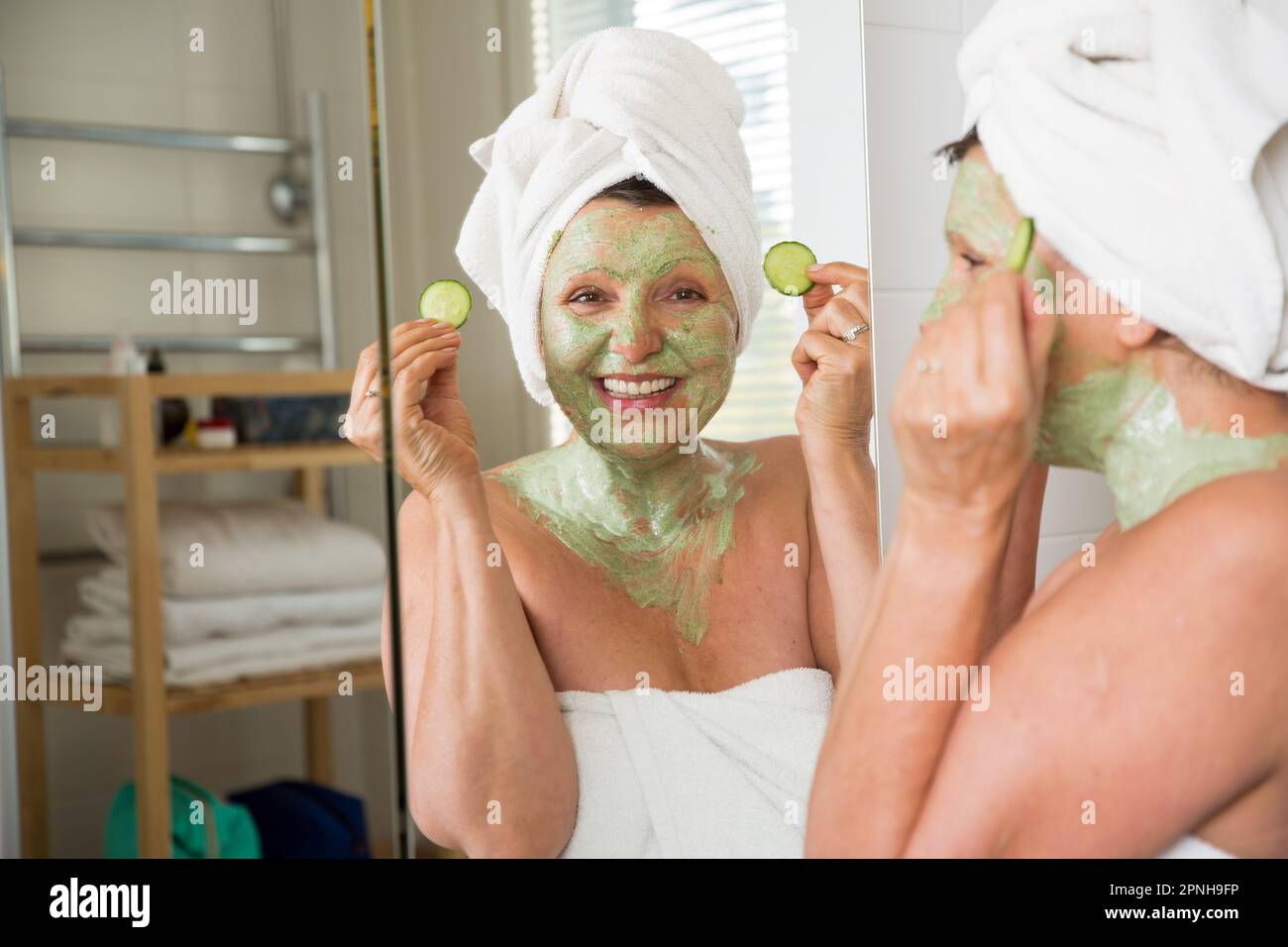 Mature beautiful woman applying green homemade DIY facial mask on skin. Looking in the mirror in bathroom, having fun. Wrapped in a towel. Skincare Stock Photo