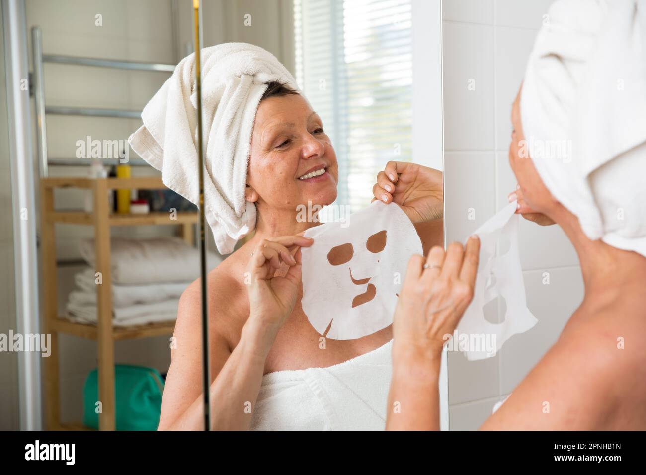 Mature beautiful woman applying facial sheet mask on skin. Looking in the mirror in bathroom, Wrapped in a towel. Skincare routine and anti-aging Stock Photo
