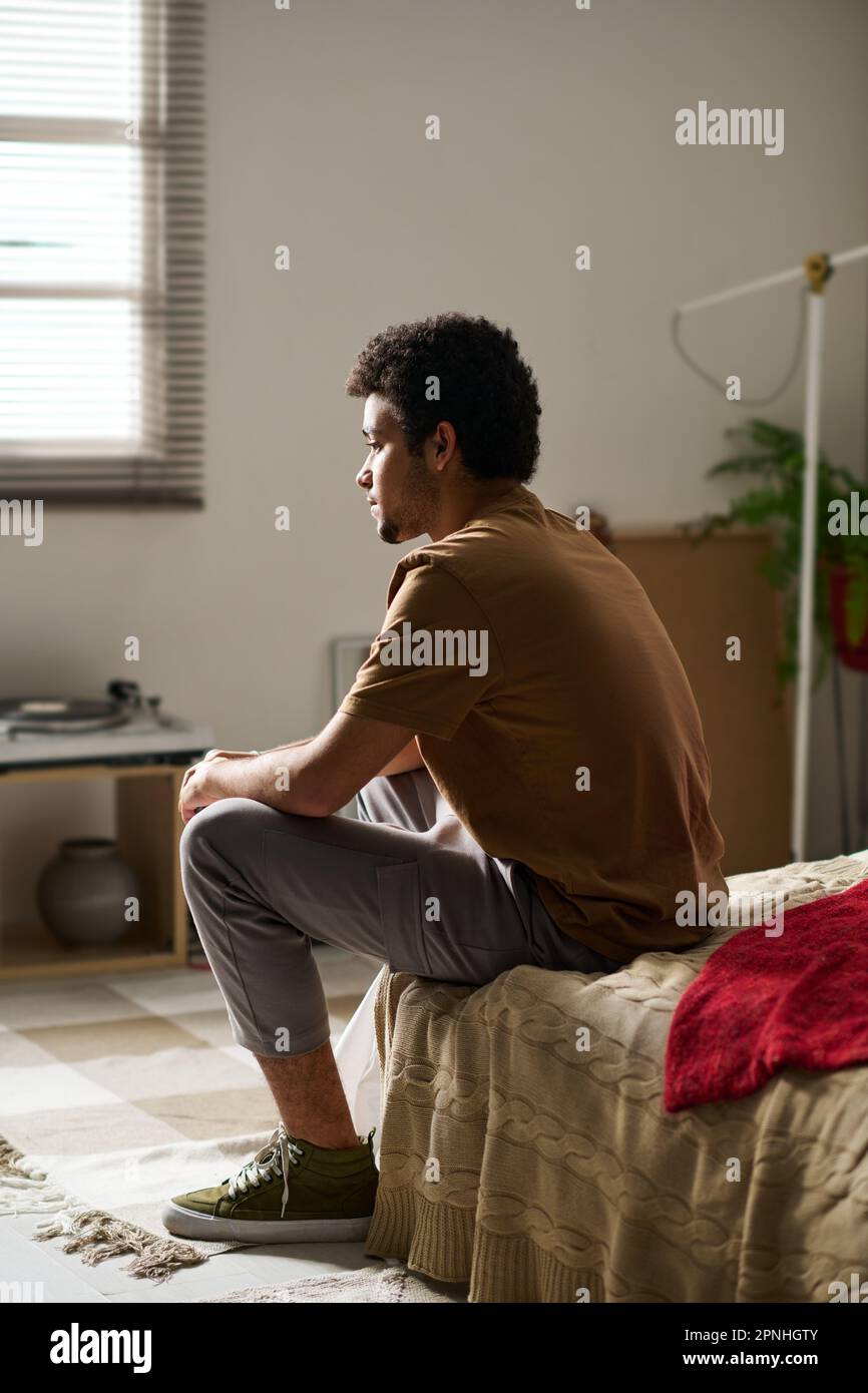 Rear view of young guy sitting on bed in thoughts while staying alone ...