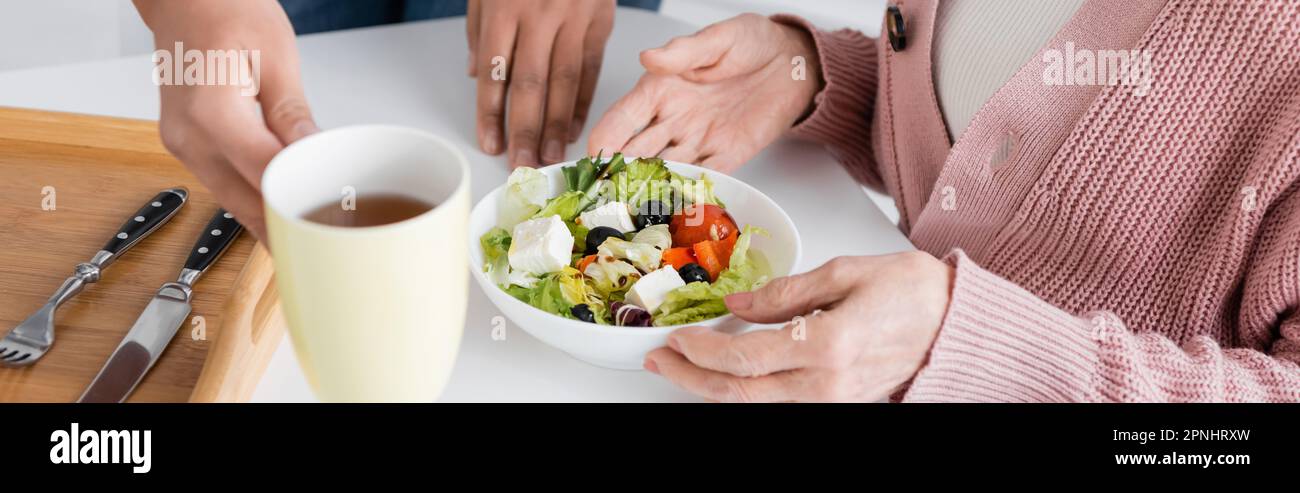 cropped view of multiracial social worker serving lunch near senior woman, banner Stock Photo