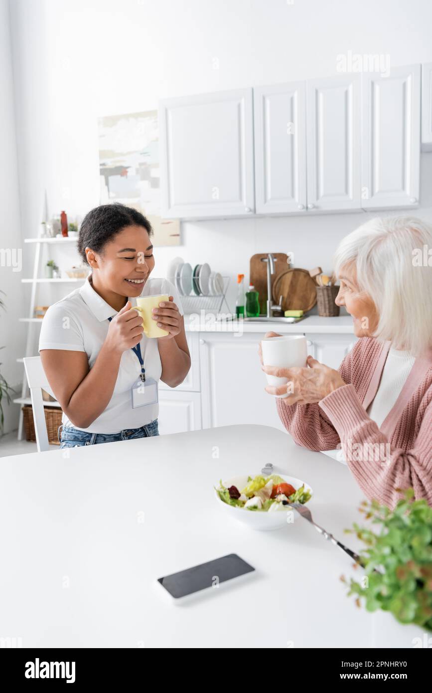 happy multiracial social worker having tea with positive senior woman in kitchen Stock Photo