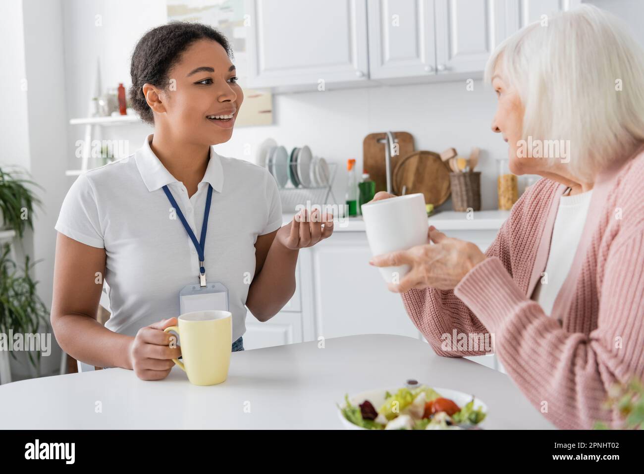 happy multiracial social worker having conversation with senior woman over cups of tea Stock Photo