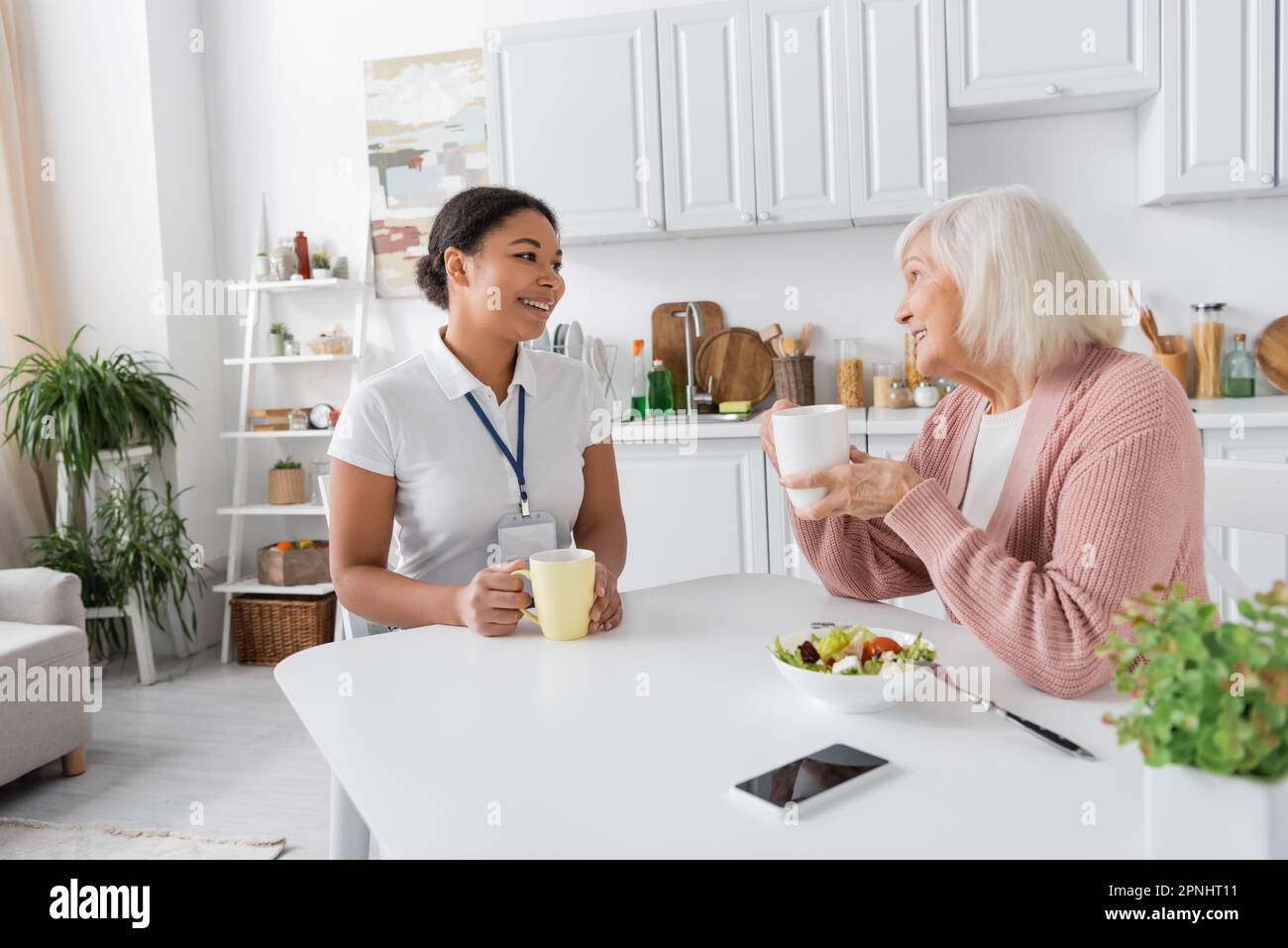 happy multiracial social worker having tea with senior woman in kitchen Stock Photo