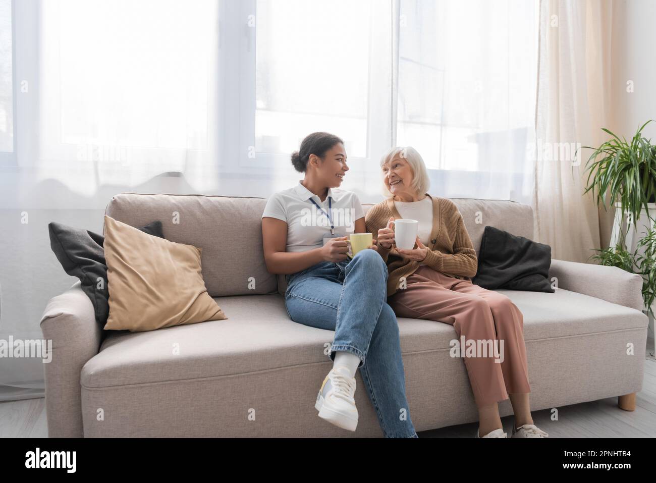 happy multiracial social worker having tea with senior woman in living room Stock Photo